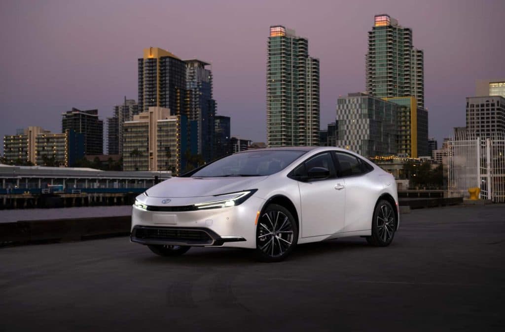 A white 2024 Toyota Prius, fifth-generation XW60, parked on a waterfront promenade with a city skyline in the background